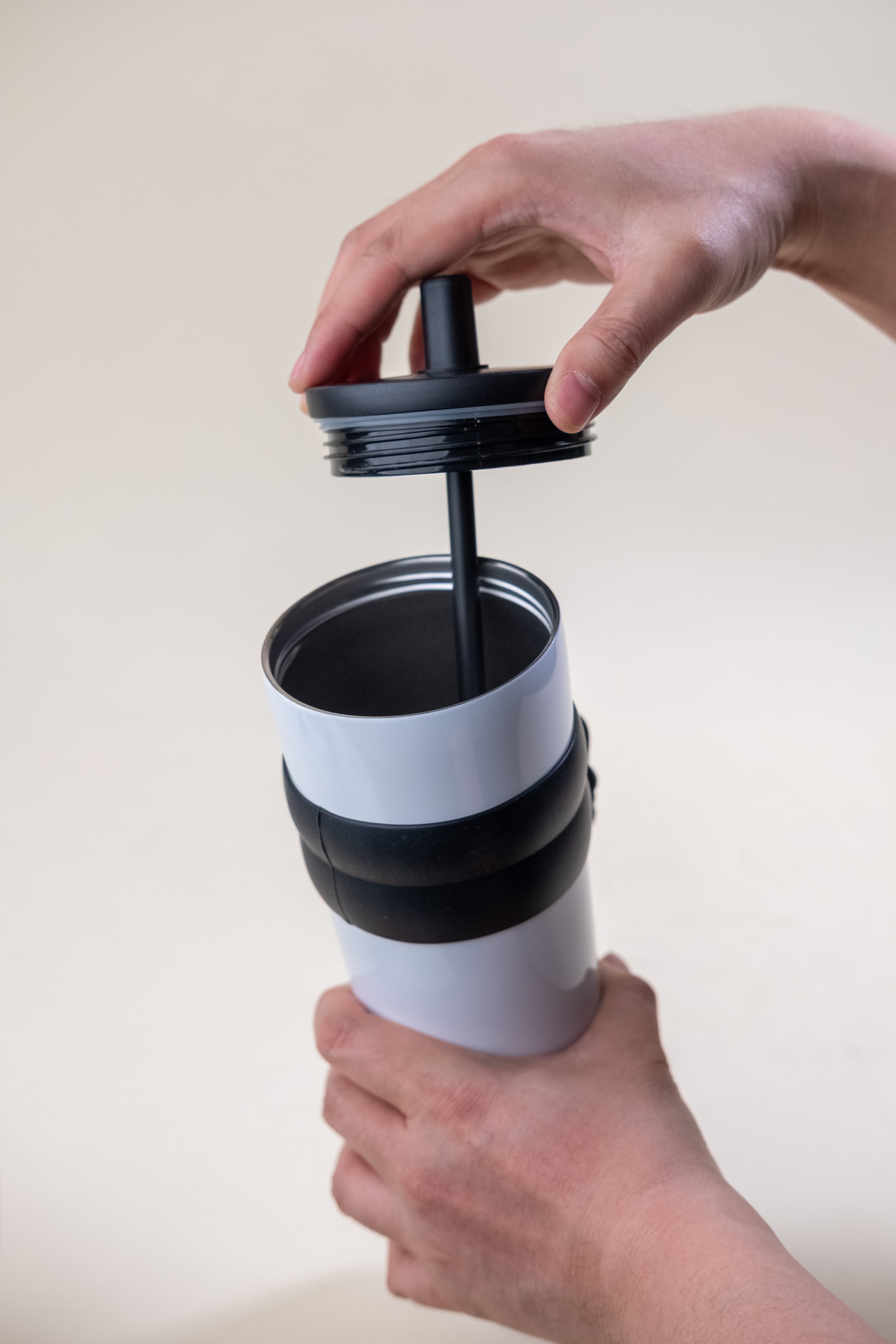 A person's hands opening the lid of a white and black stainless steel straw tumbler, showcasing the tumbler's rubber grip and design focused on user-friendly access.