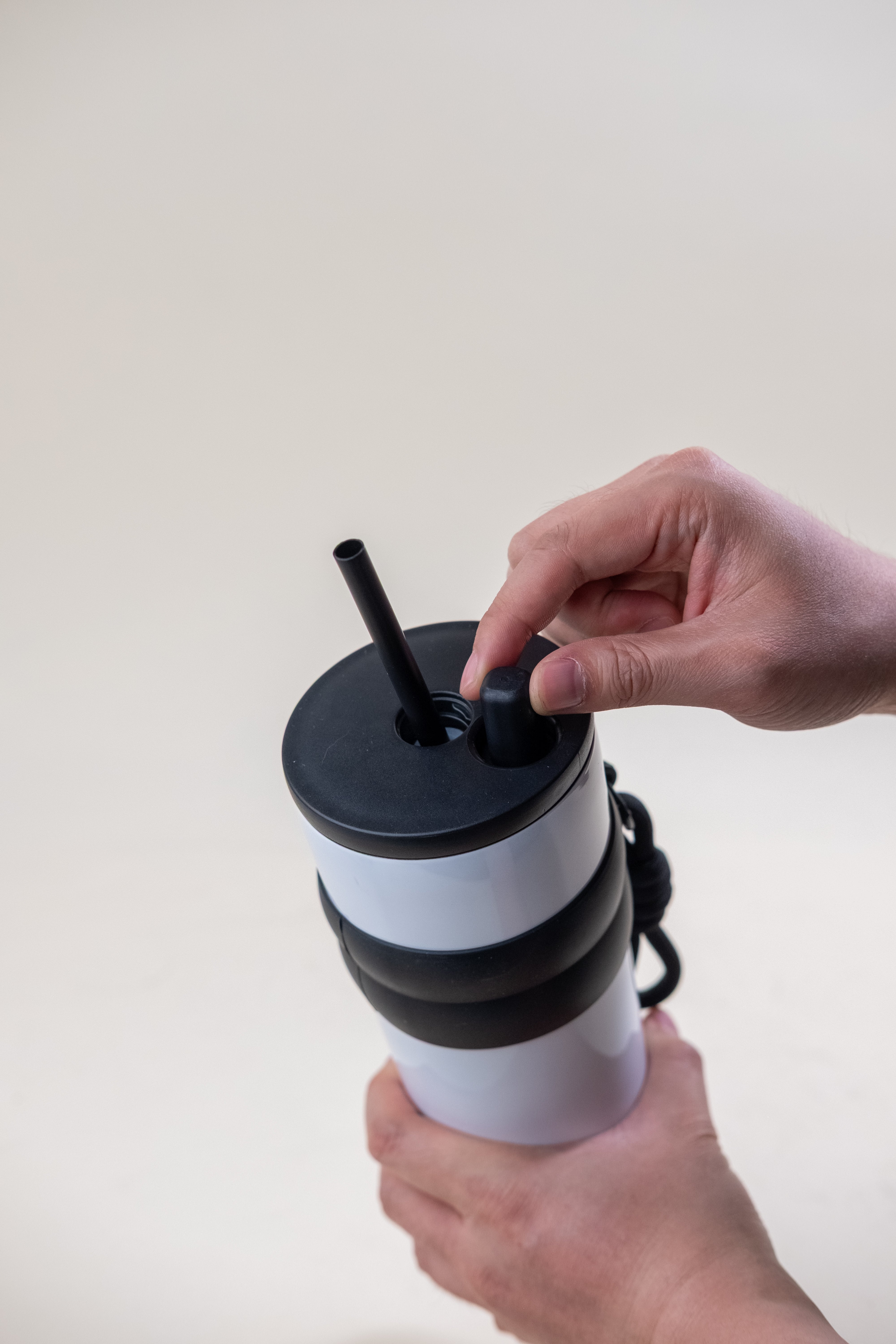 A close-up view of a person's hand placing a straw into the lid of a white and black stainless steel straw tumbler, emphasizing the tumbler’s convenient design and ease of use.