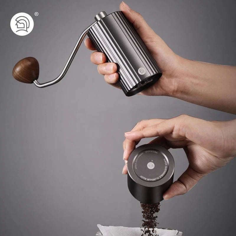 Manual coffee grinder with a ribbed black body, stainless steel crank, and wooden handle, pouring freshly ground coffee into a filter