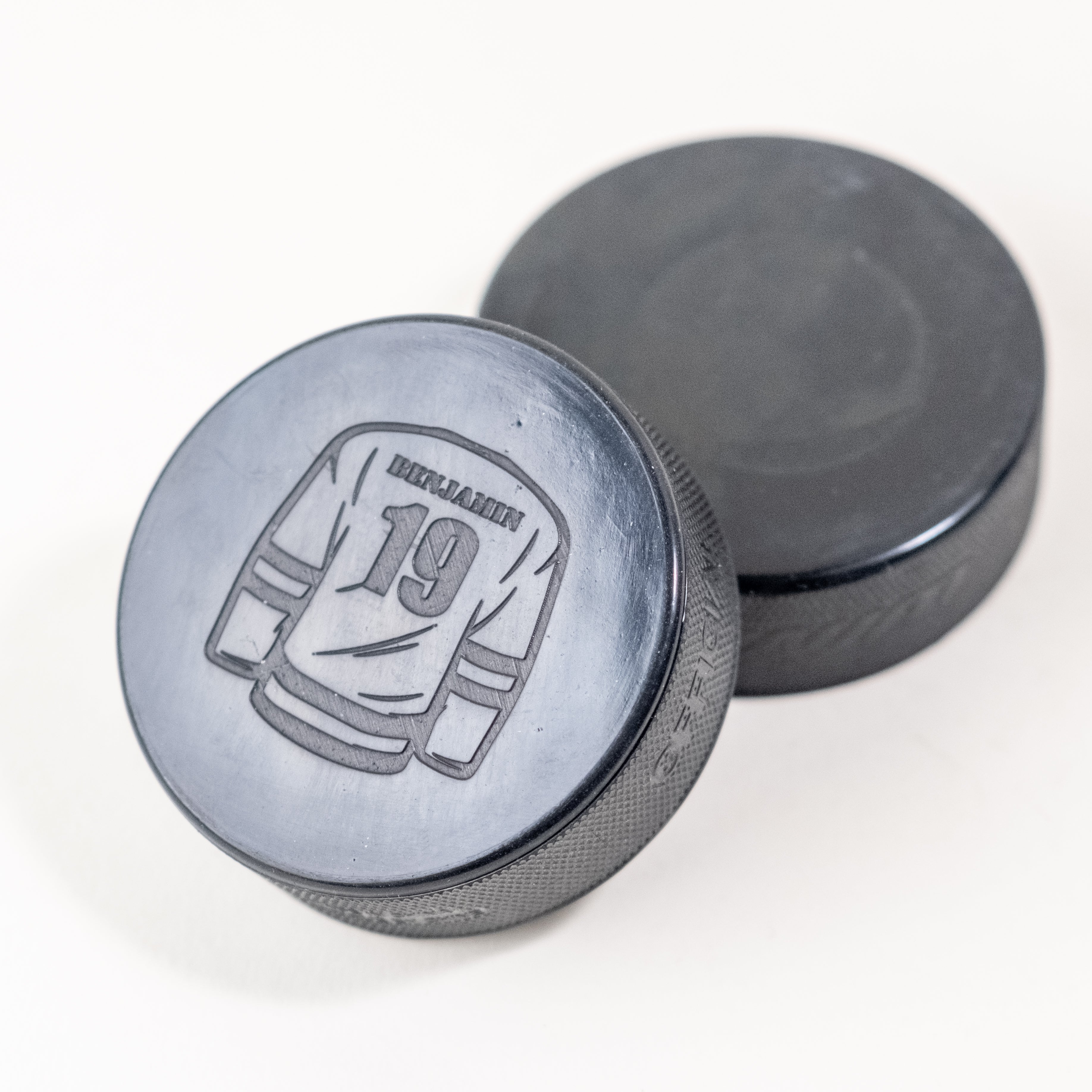 Custom hockey puck with an engraved jersey design featuring the name 'Benjamin' and number '19', placed alongside a plain black puck.
