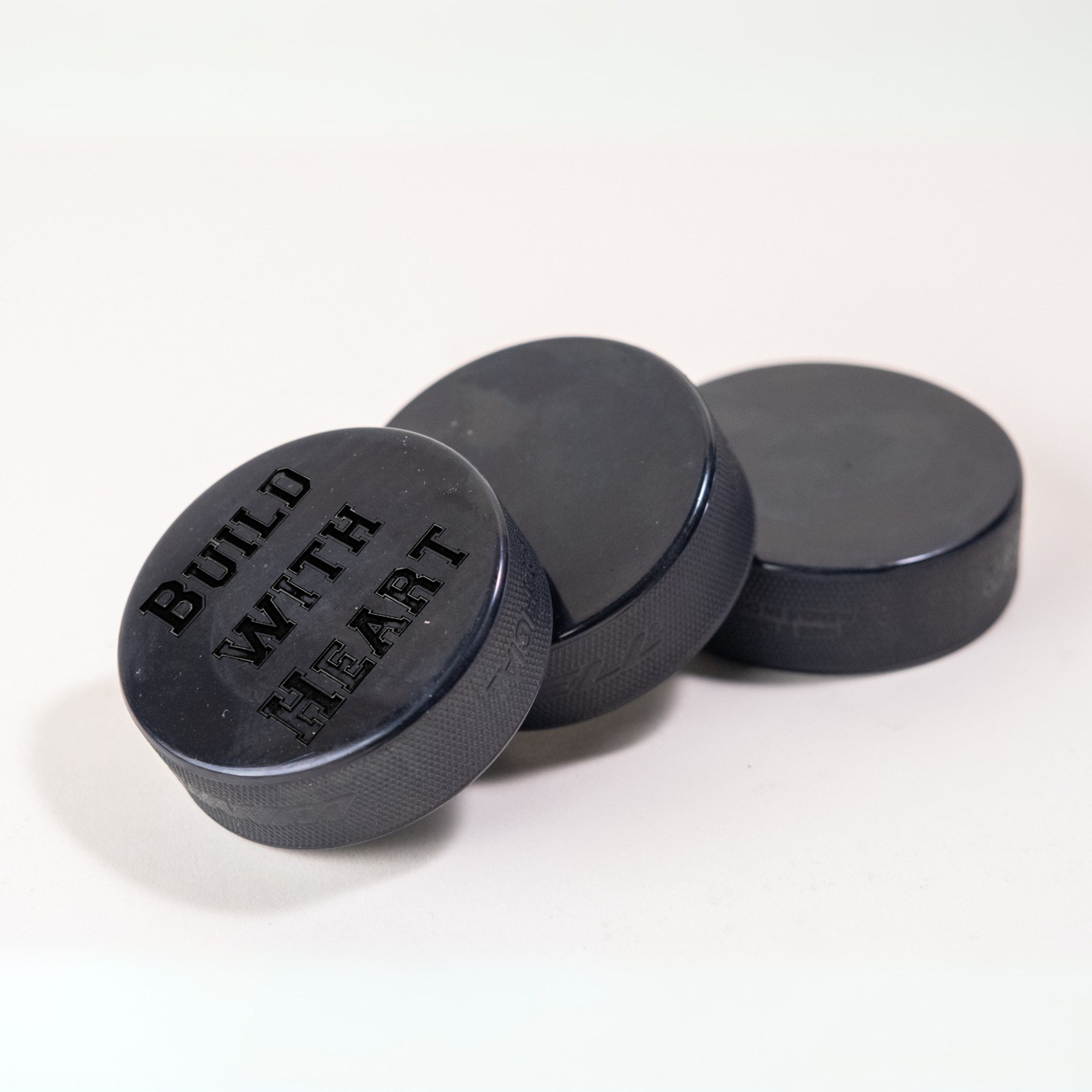 Custom black hockey puck engraved with 'BUILD WITH HEART' alongside two plain pucks in the background.
