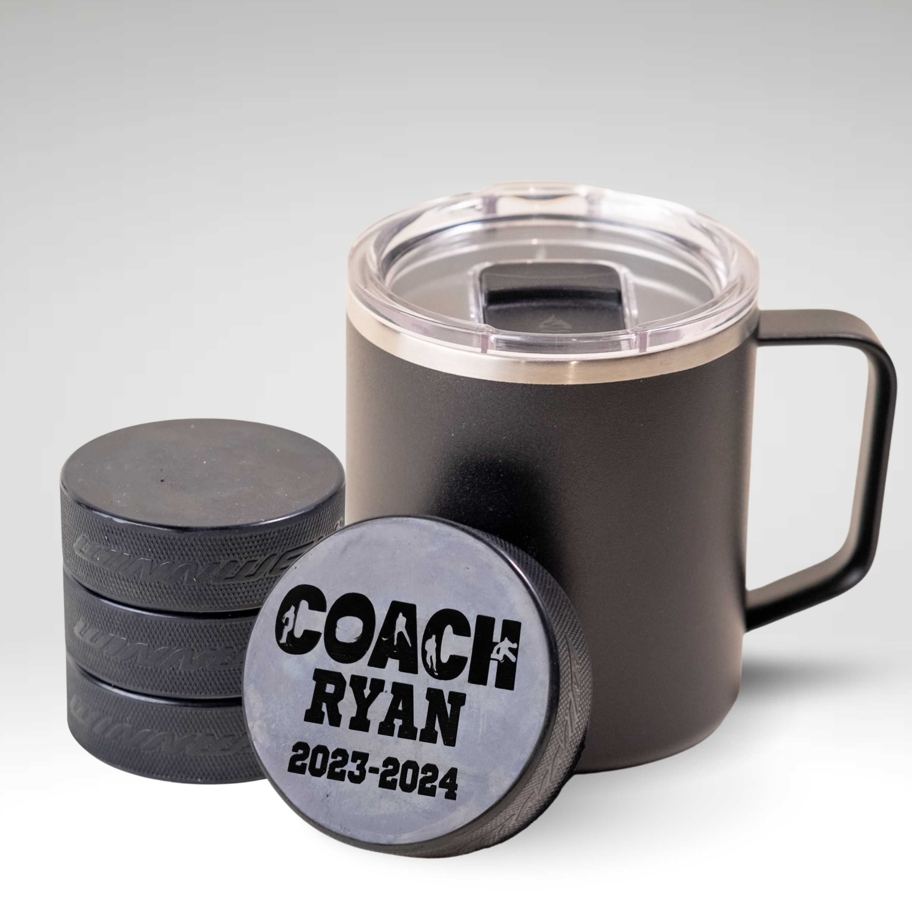 Custom engraved hockey puck for Coach Ryan 2023-2024, displayed with a black insulated coffee mug and stacked pucks.