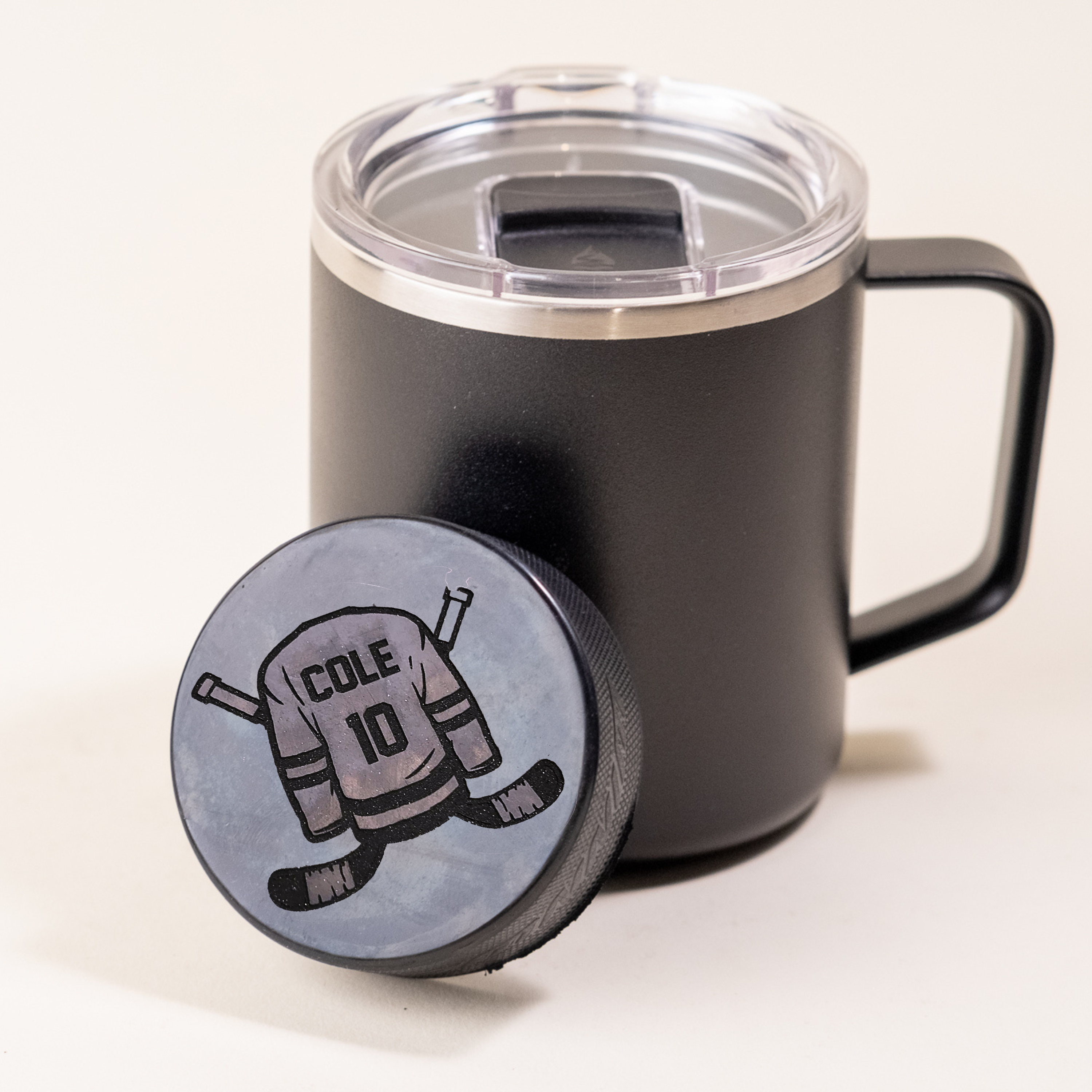 Custom hockey puck featuring a jersey design with the name 'Cole' and number '10', placed in front of a black insulated tumbler with a clear lid.
