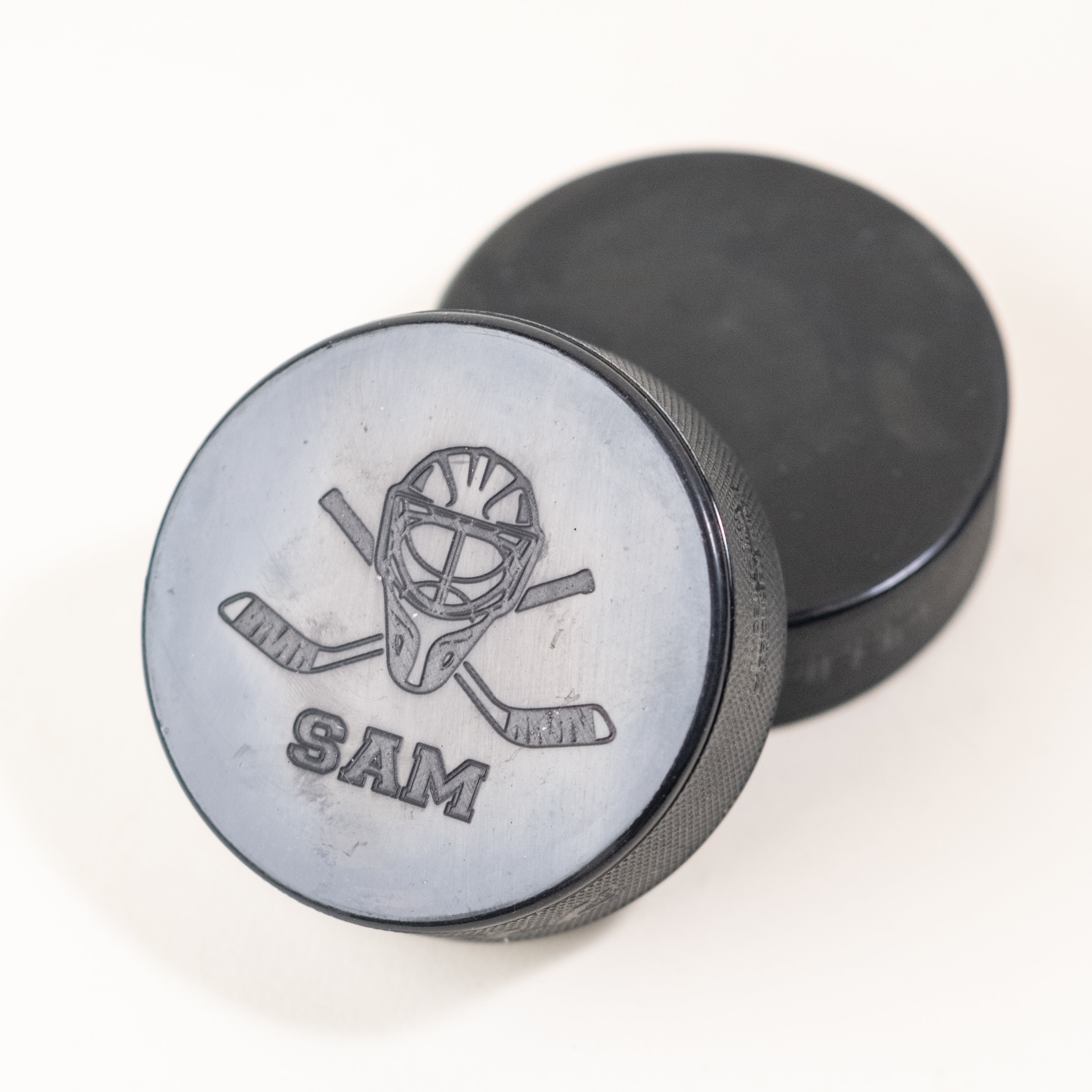 Custom hockey puck featuring an engraved goalie mask design with crossed sticks and the name 'SAM' below it, placed alongside a plain black puck.