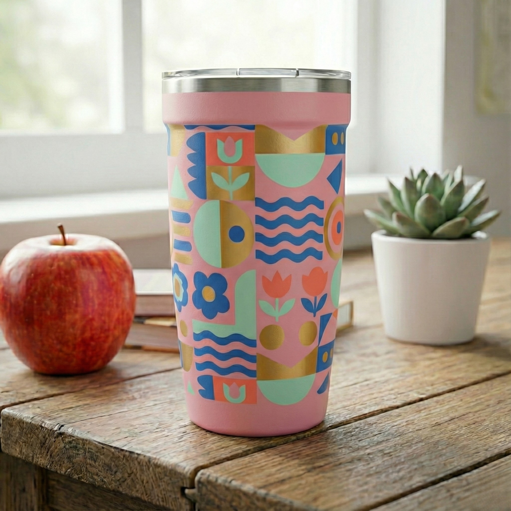 Light pink Peyto 16oz travel cup featuring a blue, green, and gold geometric floral pattern sitting on a wooden table next to a succulent and apple.