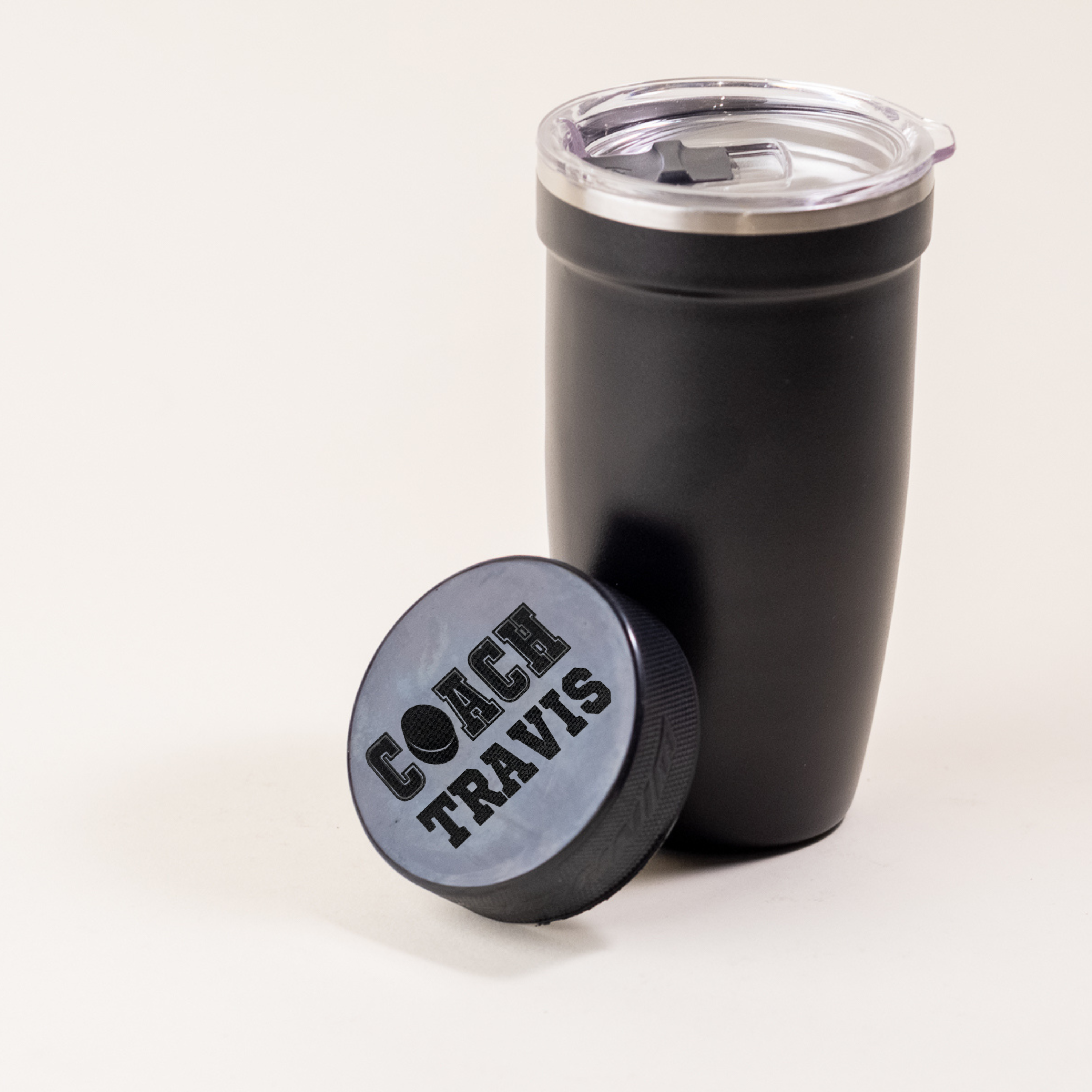 Custom engraved hockey puck with 'Coach Travis' design next to a black insulated tumbler.