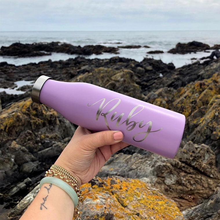 LAMOSE personalized lavender matte stainless steel bottle held by hand outdoors, custom engraved with 'Ruby', rocky ocean backdrop.