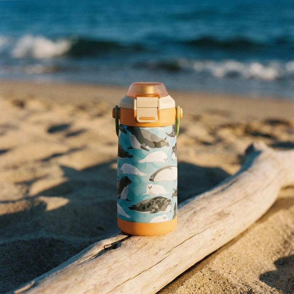 Lifestyle shot - LAMOSE Goose 14oz kids water bottle featuring a light blue body with a pattern of gray and white cartoon seals, orange bottom bumper, and green handle.