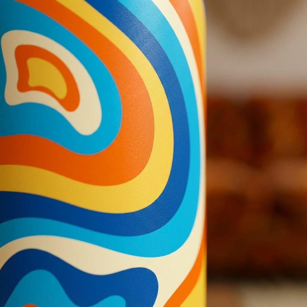 Detail shot - Grouse 12 oz insulated tumbler featuring a retro 1970s psychedelic swirl pattern in orange, blue, and cream on a teak table.