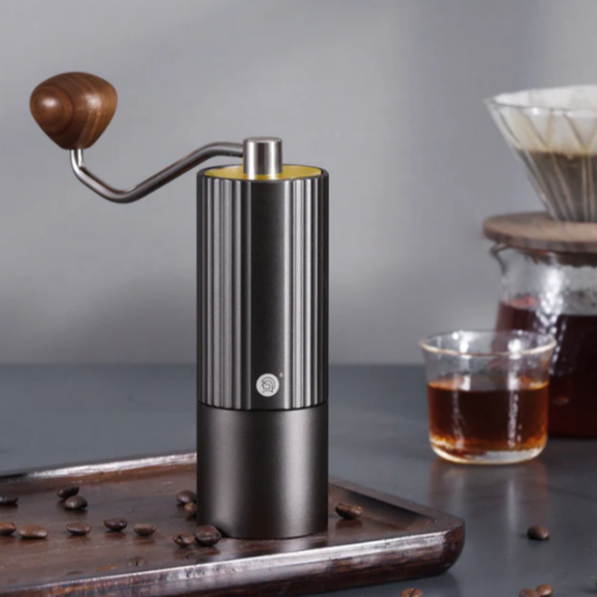 Elegant manual coffee grinder with a black ribbed body and wooden handle, placed on a wooden tray with coffee beans, alongside a glass of freshly brewed coffee