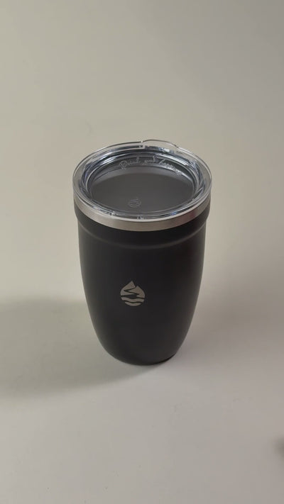 Hand holding an open black insulated tumbler with a stainless steel interior and a removed clear spill-resistant lid. Double-wall vacuum design for hot and cold beverages.