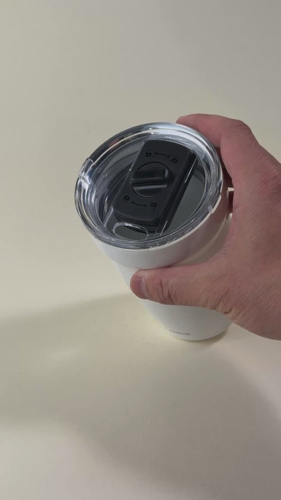 Hand adjusting the sliding lock mechanism on a white insulated tumbler with a clear spill-resistant lid. Stainless steel travel mug designed for hot and cold beverages.