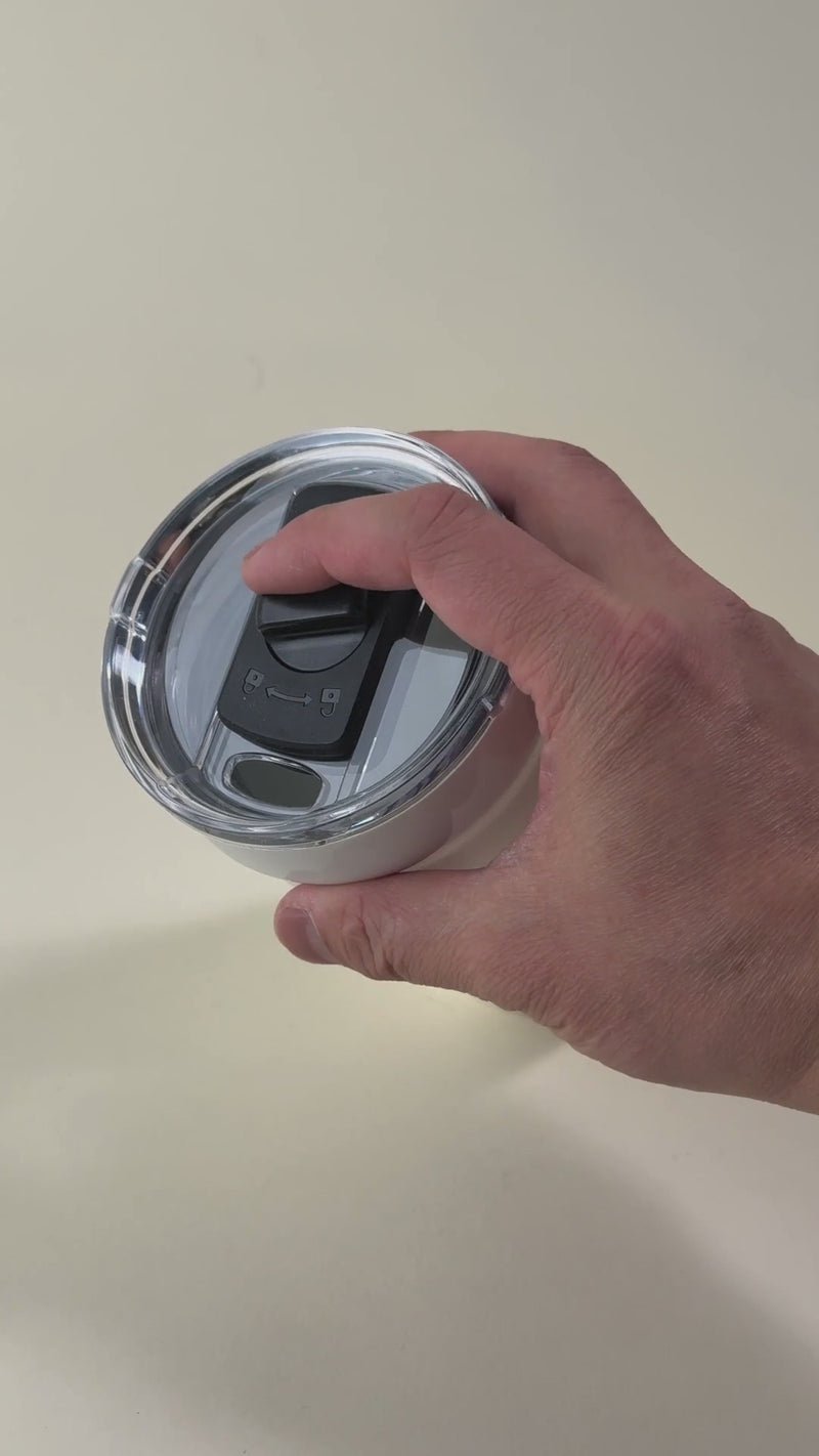 Hand pressing the sliding lock on a white insulated tumbler with a spill-resistant lid. Stainless steel travel mug designed for secure and convenient drinking.
