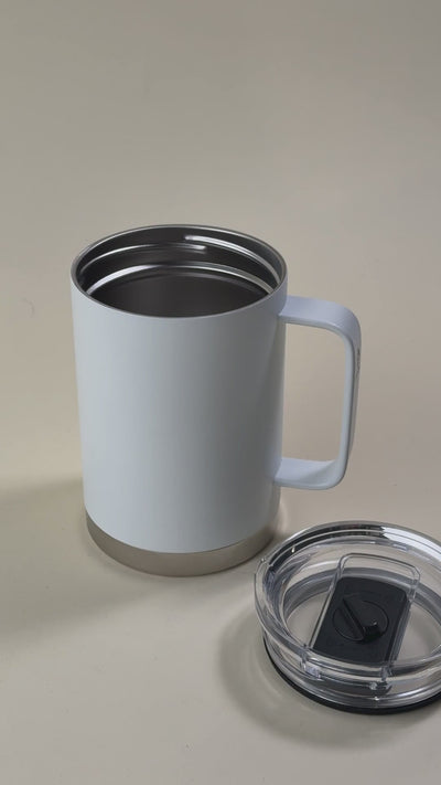 White insulated stainless steel mug with a sturdy handle, held by a hand, alongside a transparent spill-proof lid.